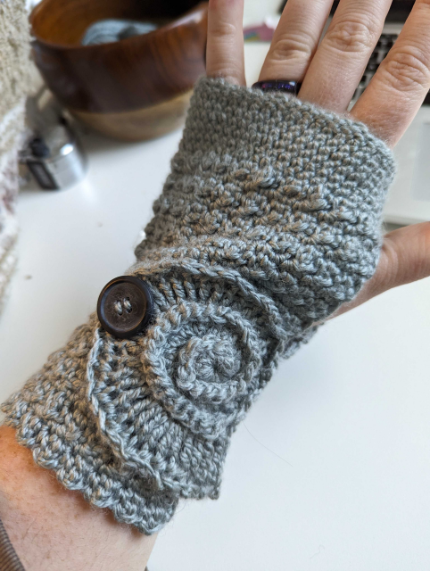 A gray wrist warmer on a human wrist. The wrist warmer is made from crocheted yarn. The design of the stitches forms a ammonite shape on the wrist.