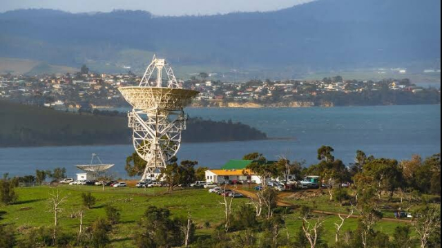 A large radio telescope sits atop a grassy hill, with a wide river in the background. Beside it and down the hill a bit sits a smaller dish. This is the Mount Pleasant Radio Observatory outside Hobart in Tasmania.