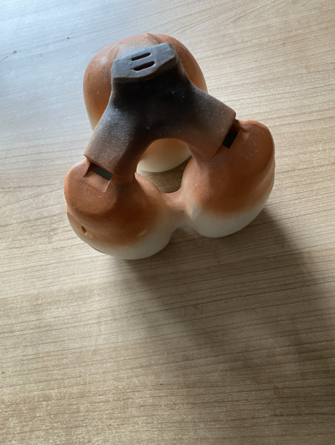 The huaca sitting on my coffee table. Its three globular chambers, two equally-size ones at the front and a larger at the back are connected at a top via the Y-shaped configuration of air ducts radiating out from the mouthpiece. The chambers are also connected to each other by horizontal extensions at their widest part, and highlighting their triangular configuration. The mouthpiece has two slits; the front one for the two melody chambers and the back for the drone. The front chambers both have 3 finger and 1 thumb holes, and the drone chamber has two palm holes; one for either hand. The chambers have flat bases, allowing you to support the entire flute on your pinkies. In short, it's quite a lot to keep track of. The flute is decorated with clay of various natural colours.
Because the shape is rather abstract, when I've thrown this photo into AI it has resulted in hilarious descriptions of it somehow thinking it's a dog toy for dispensing treats, a stand of some kind, a massager, or a funny statue of a cow. All EPIC fails, but just too funny!