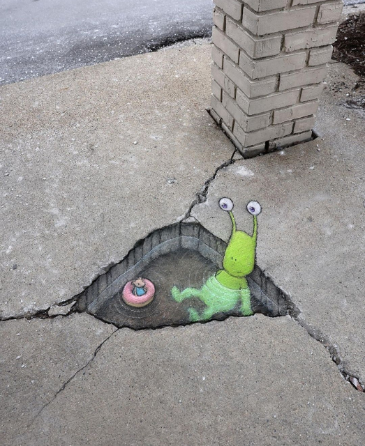 Streetart. In a painted pothole in a beige concrete slab path with cracks, next to a small brick pillar, a 3D image of a mouse together with an alien was drawn in chalk. A puddle of water was masterfully drawn into the hole with chalk. The mouse in the blue dress (Nadine) swims in it in a pink rubber boat. Next to her, an alien three times her size sits in the water and looks at two antennae with its stalk eyes.
Title: "Nadine and the unexpected bath 
Day Companion (with a guest appearance by Sluggo)"