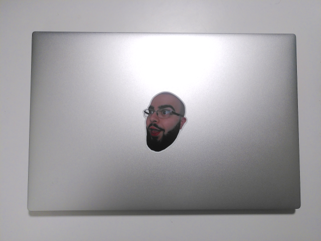 A top down photo of a silver laptop lid, with a single sticker of someone's face in a slightly shocked, slightly silly expression. 