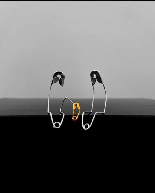 Photography. A color photo of three safety pins with a family connection. Three safety pins have been positioned on a black surface against a gray background. To the right and left are two curved silver safety pins and in the center is a small gold one. The pins are bent so that they appear to be sitting next to each other. The left safety pin is bent with its point towards the small golden safety pin, as if hugging it. The three look like mom, dad and child.
Title: "Adopted child"