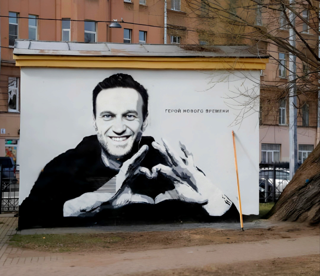 Photography. A color photo of a plastered wall of a substation on April 28, 2021 in St. Petersburg, Russia. The white poster shows an artistically edited black and white photo of Russian opposition activist Alexej Nawalny from the front. He is forming a heart with the fingers of both hands. Next to it is written in Russian "Hero of a new era". A long yellow stick leans against the billboard. (Brown house fronts can be seen behind it. In front of it is a paved sidewalk)
Info: The billboard was pasted by unknown persons in the middle of Russia in April 2021 and painted over directly with yellow paint by the employees there. However, the photographer captured everything in photos. 
