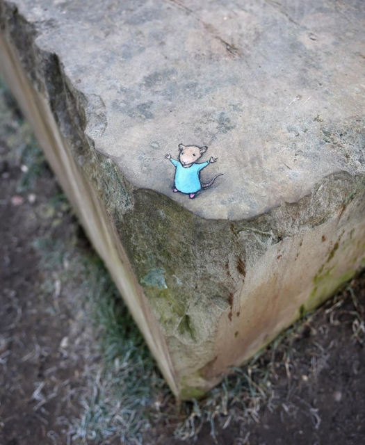 Streetart. A cute mouse with a 3D effect has been painted with chalk on a gray concrete block with a broken corner. The little mouse, whose name is Nadine, is wearing a pretty blue dress and is standing on top of the edge of the stone, stretching her arms in the air. (There is dark sand and some grass on the ground).
Title: "Nadine embraces life on the edge"