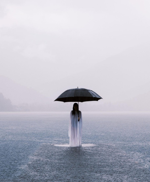Photography. A color photograph
of a young woman standing in a lake. The photo is bathed in light colors such as gray and light blue. In the center of the picture, a young woman with long black hair in a long white robe and a black umbrella is standing in a lake. Her face is unrecognizable. Gray-blue water ripples around her. Sunk in the mist, a mountain landscape can be seen on the horizon.
Info: The young woman is standing on a wooden walkway that is completely submerged in water. Zhang Ahuei is a photographer based in Taiwan. He focuses on portrait and fashion photography. His mostly female characters move between the real and the surreal as if in a daydream.