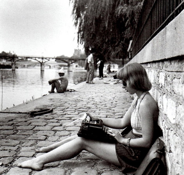 Vintage black and white photography.

Woman sitting at the bank of a river in a city.

She has a mechanical typewriter on her lap.
