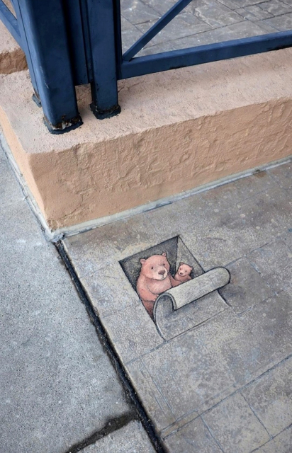 Streetart. Two cute bears have been painted with chalk and a 3D effect on a gray sidewalk. The sidewalk consists of rectangular gray slabs, and a "hole" has been painted on one of them. A rolled-up "stone" has been painted over the painted hole. It looks like a sardine tin. Now you can look inside and see... two bears looking out. A big bear shows a little bear the world outside.
(The photo also shows an orange-painted concrete edge with blue metal pipes)