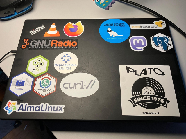 A ThinkPad laptop covered in various stickers from tech and software companies/projects, including logos like the Firefox browser, GNU Radio, and Wikimedia, alongside other miscellaneous stickers.