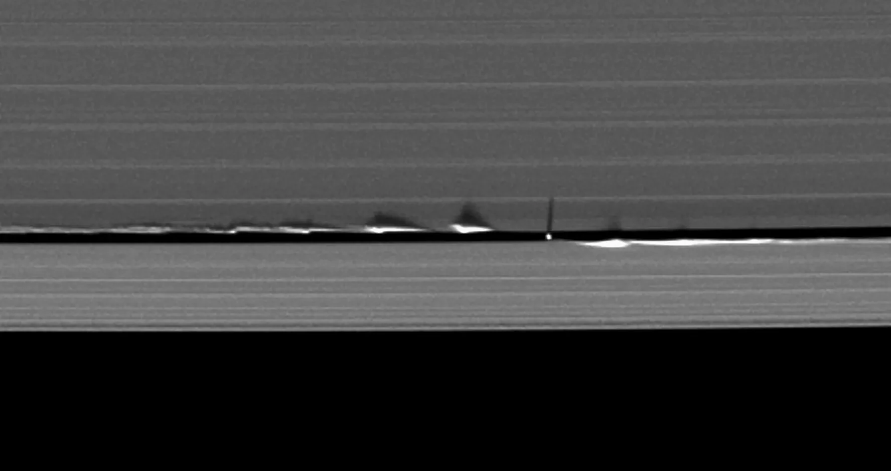 The bottom third of the image is filled with darkness. The top two thirds show the rings of saturn, forming patterned density waves vertically. In the center of the image there is a gap in the rings, and the moon Daphnis can be seen sweeping out a path. Both in front of and behind the moon, waves of ring dust are being created in a plane perpendicular to the camera and the rest of the ring structures.