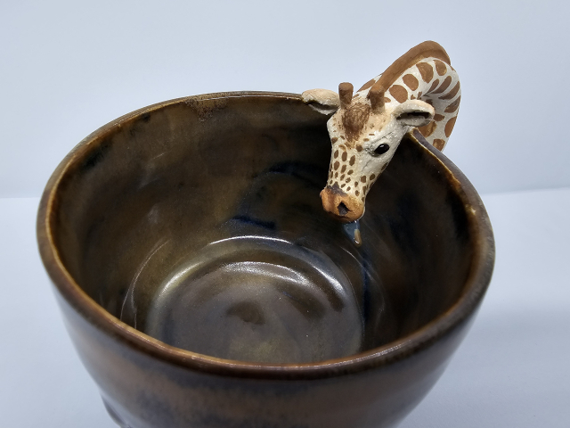 View of giraffe head entering mug with its tongue sticking out. The tongue is blue but has a splodge of brown glaze on it as though it's just licked the inside of the mug 