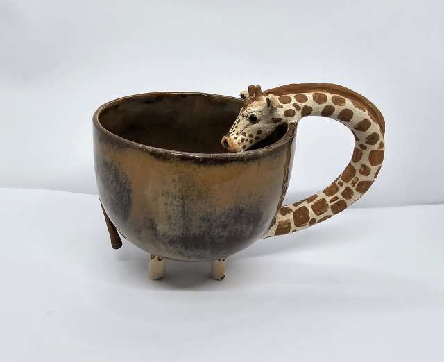 Side view of giraffe mug. To the left is the tail, 4 legs underneath and the giraffes neck form the handle with its head sticking in to the body of the mug. The mug is brown ish 