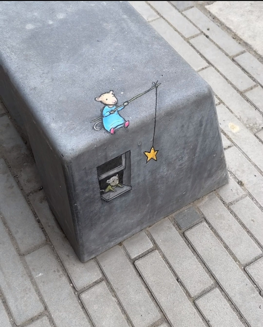 Streetart. Two cute little mice were painted with chalk and a 3D effect on a dark gray, low concrete bench. At the top of the bench sits a mouse in a blue dress and pink shoes (Nadine), holding a fishing rod with a golden star. The star is above a painted window on the side of the bench. Another mouse is looking out of it. What will she dream? Title: "Nadine and the Wishing Star"