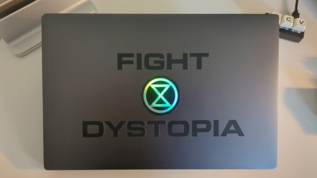 An overhead shot of a gunmetal grey laptop lid. There is a round sticker in the middle with a simplified reflective hourglass over a black background. This sticker is framed by the words 'Fight Dystopia' above and below. In the upper right hand corner of the photo the C and V keys of a Stack Overflow keyboard can be seen.