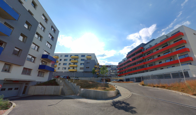 Foto nově vzniklého obytného souboru v Plzni. Bytovky sice perfektně splňují parkovací minima, požadavky na proslunění bytů, všude je dostatek prostoru pro vytočení hasičského auta nebo průjezd IZS, ale místo je hnusné. Řešení chodníků je podřízené vedení asfaltové vozovky, v lokalitě je minimum zeleně, ulice nemá lidské měřítko, přízemí domů schází obchodní parter.