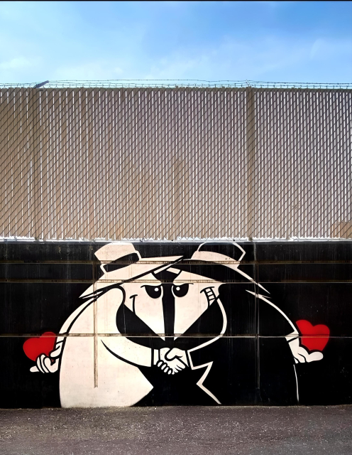 Streetartwall. A cute comic-style mural was sprayed on a low wall with a fence above it, which belongs to an industrial company. The background is painted black. Two mice or rats with pointed faces are painted on it. The mouse on the right has a white face and is wearing a black suit and hat. He is grinning and shaking hands with a smiling mouse in a white suit and hat. Both are hiding a red heart behind their backs. The two look like mafia gangsters from the 1950s, but instead of revolvers they have a lot of love with them.
