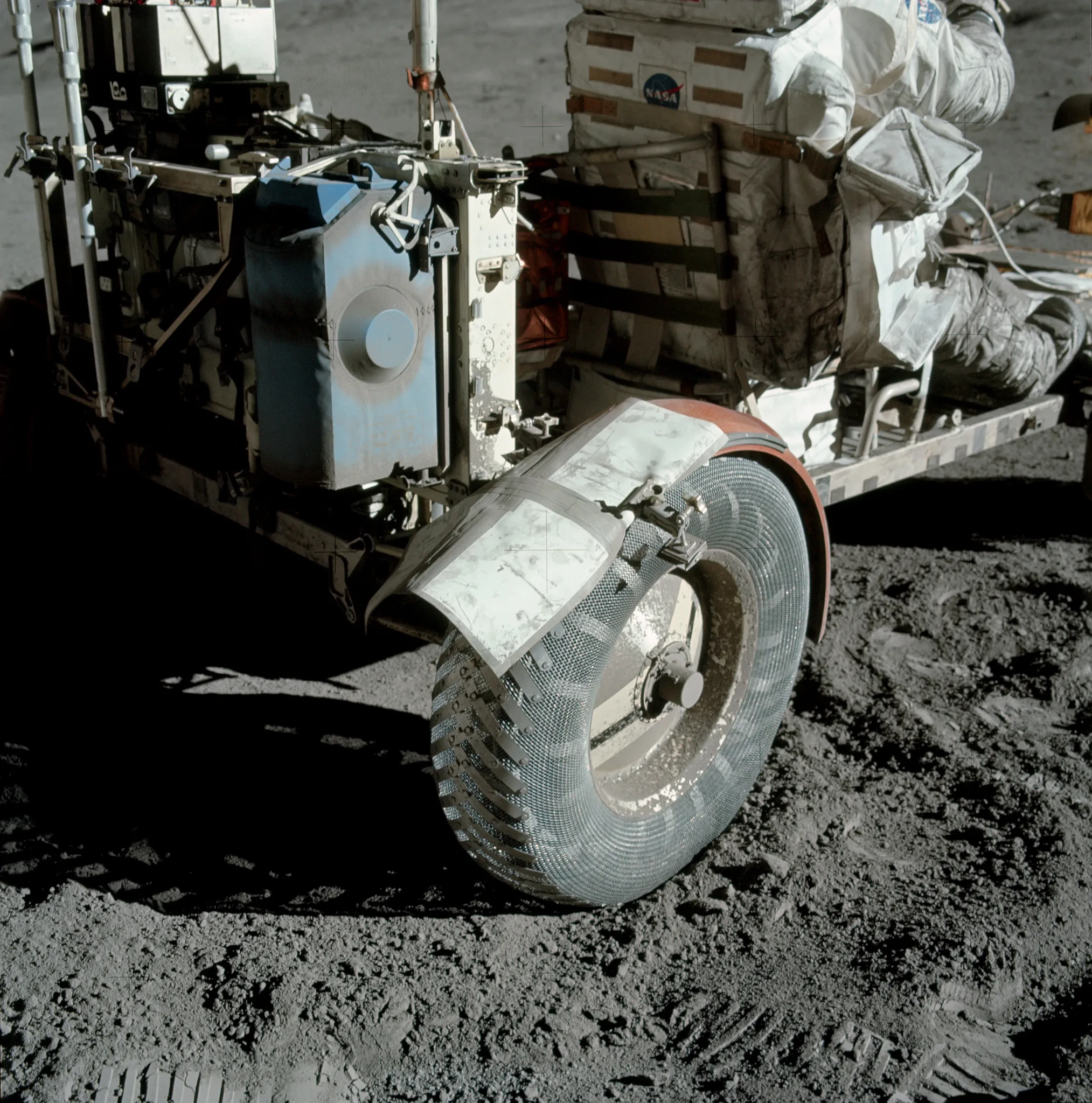 A photo of the lunar rover vehicle taken from behind, centered on the right rear wheel. A replacement fender is haphazardly attached. A dust-coated originally blue box is mounted above the wheel, and a suited up astronaut is driving the vehicle. The astronauts suit is also covered in lunar dust. https://www.nasa.gov/history/alsj/picture.html