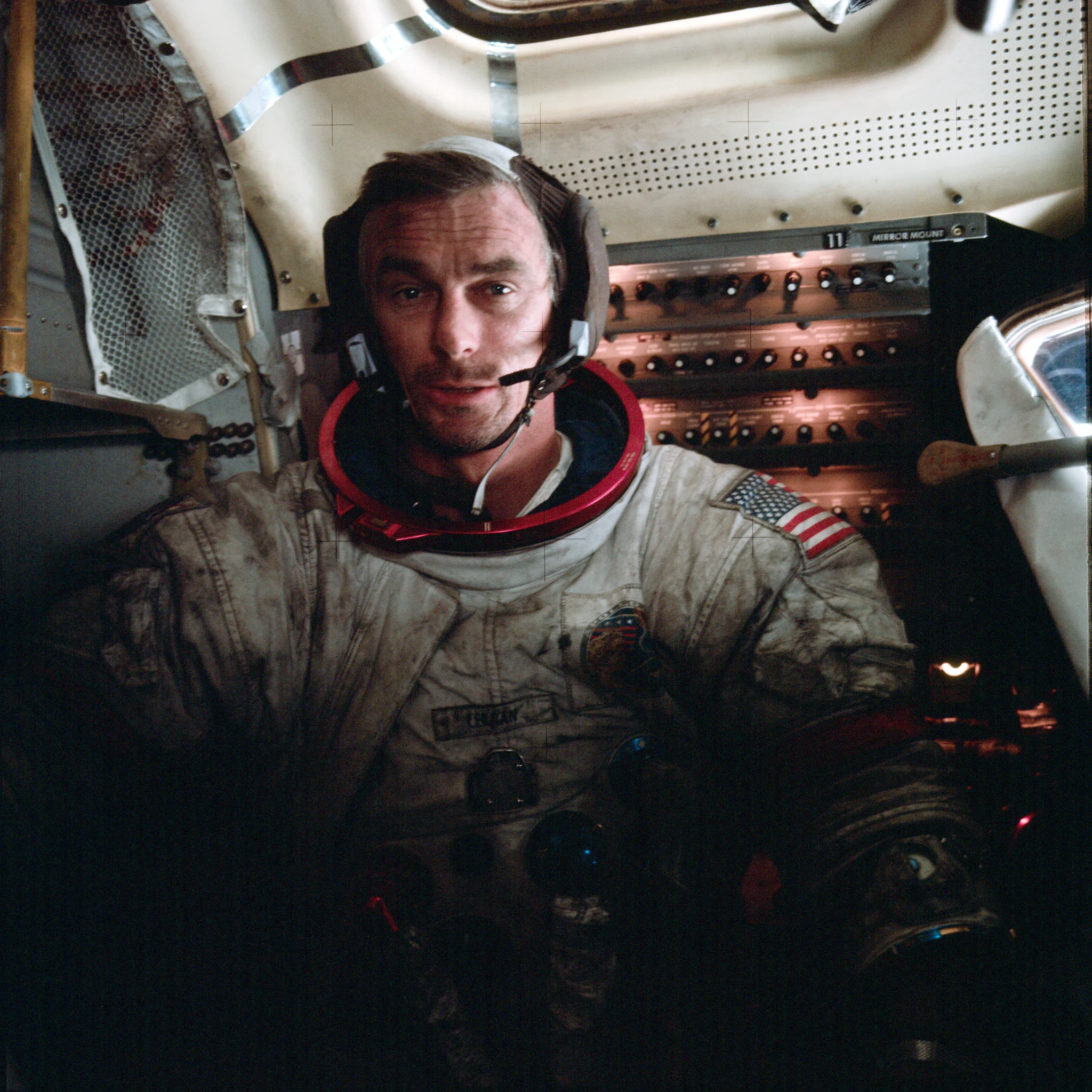 Astronaut Gene Cernan sits in his spacesuit, helmet off, inside the Apollo 17 module. Various knobs, tubes, and buttons can be seen behind him. His suit is completely covered in black lunar dust. https://en.wikipedia.org/wiki/Lunar_soil#cite_note-16