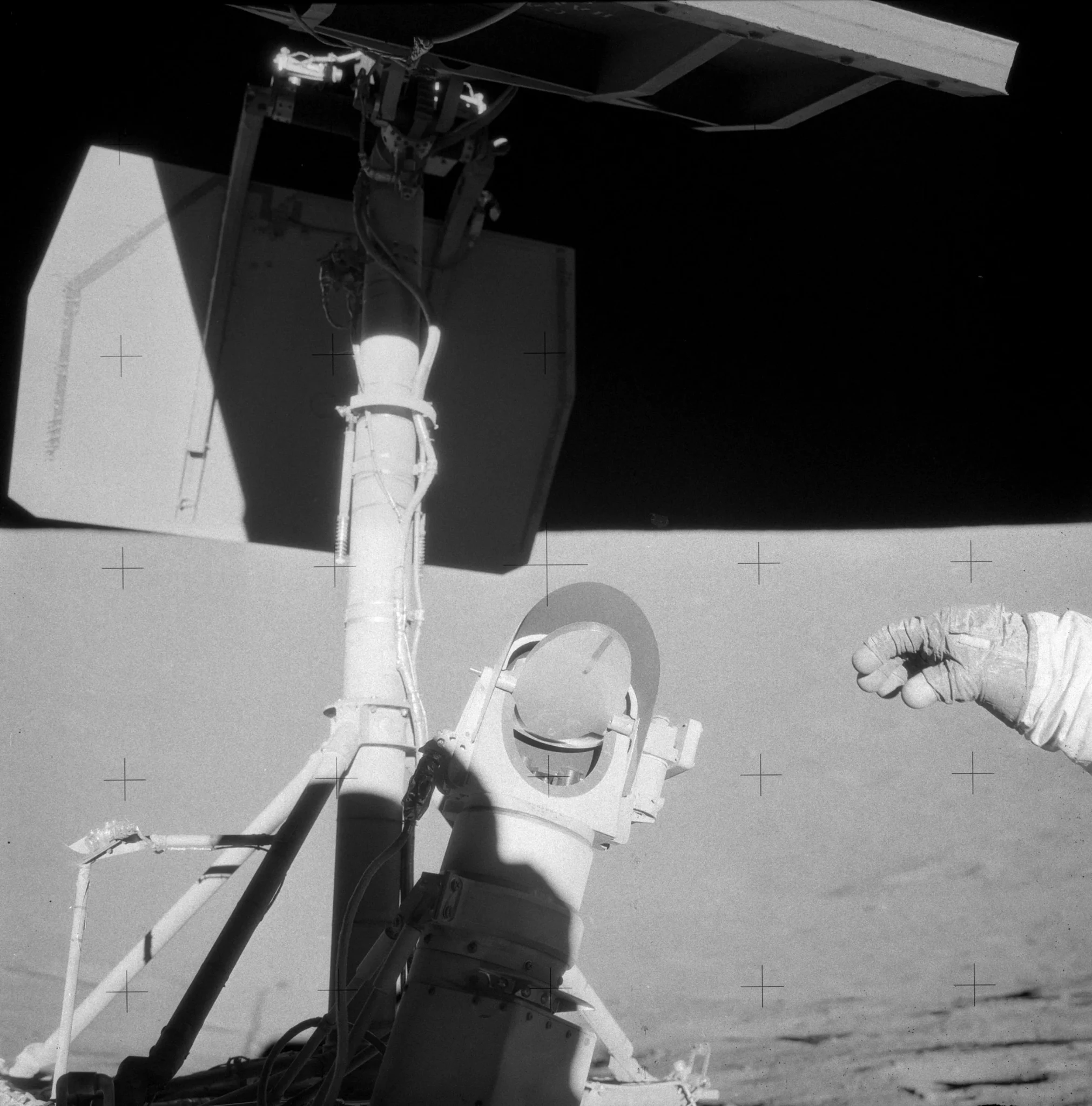 A large vertical pole holds metal panels aloft to the left of frame. In the center, a scientific instrument with a mirror covered in dust has had a single finger drawn across it, showing just how dusty it is. To the right of frame is an astronaut’s outstretched hand. https://www.nasa.gov/history/alsj/picture.html