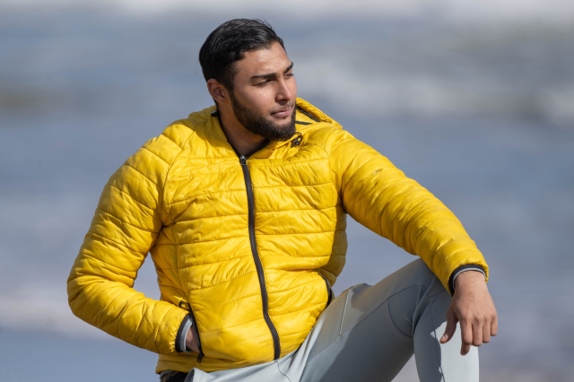 Handsome dude in a yellow parka and grey pants posing on the beach.