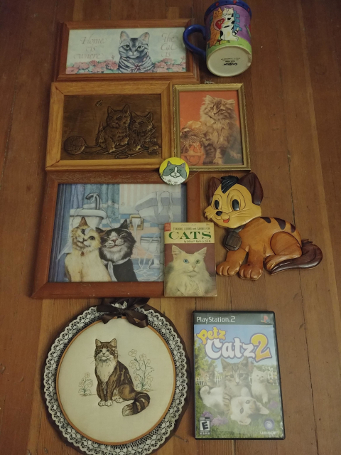 A photo of 10 cat related artifacts, laid out on a hardwood floor. Summary of items, left-to-right and top-to-bottom:

- A wooded framed watercolor of a cat with the words "Home is Where the Cat is"
- A large coffee mug, with a painted on image of two cats (visible side)
- A wooden framed, copper relief of two kittens playing with a ball of yarn
- A gold-painted, wooden framed photo of a long hair cat
- A wooden framed painting of two cats in a bathroom
- A pin of a cat
- A small booklet about cat care
- A wooden cat made of several different pieces
- A needle point of a cat on a doily 
- A PlayStation 2 game called "Petz Catz 2"