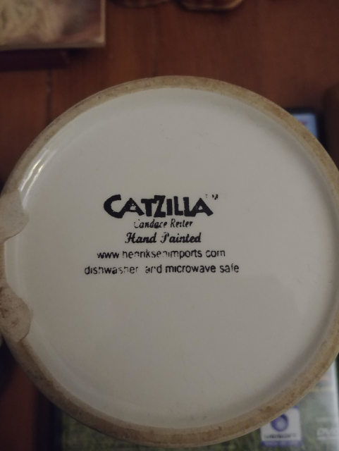 The bottom of a coffee mug. The text reads:

Catzilla (tm)
Candace Reiter
Hand Painted
www.heonksenimports.com
dishwasher and microwave safe.