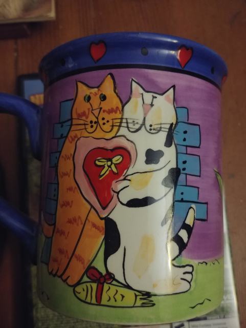 An excruciating painting of two cats together, each holding one side of a heart shaped box. Or something.

Honestly, this thing is horrific and obviously hand drawn on the mug and not a transfer.

Anyways, at their feet is a smiling fish wrapped in a bow, while behind them appears to be a fence. The cat on the left is an orange striped cat, and on the right a calico.

Or cow disguised as a calico, it's pretty rough. Also the one on the right has it's legs bent backwards in the way cat legs don't work.