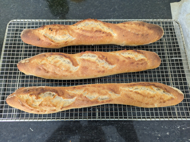Three baguettes after baking. They are golden brown and have expanded where they were scored on top. The baguettes sit on a wire cooling rack on a black marble counter top.