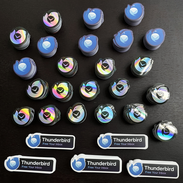 A selection of Thunderbird stickers, including the standard logo of a stylized blue bird curled around an envelope, a holographic version of the logo, and the standard logo with the text 'Thunderbird - Free Your Inbox' to the right, stand out against a black wooden surface.