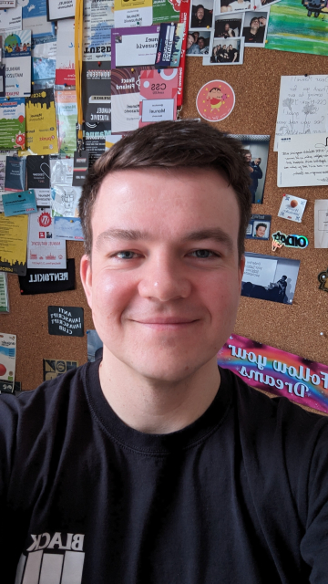 A selfie. I'm smiling, wearing a black black flag band shirt. I'm standing in front of a cork wall with passes from conferences attached to it, photos, stickers and other random stuff.
