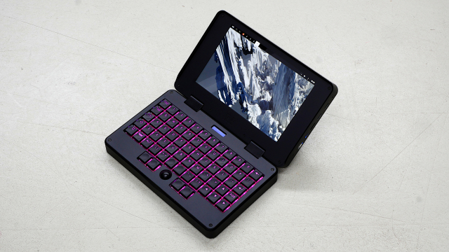 a small (roughly A5 size) black clamshell laptop with ortholinear mechanical keyboard, pink backlit keys, black mini trackball, showing mnt reform sway desktop with "mountains" background