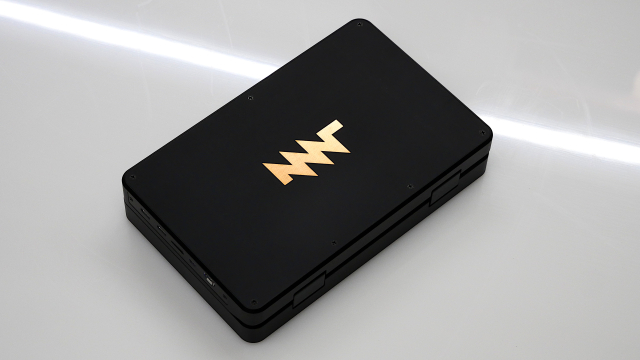 closed black mini laptop (roughly A5 sized) mnt pocket reform on shiny acrylic surface, copper mnt research logo in the middle