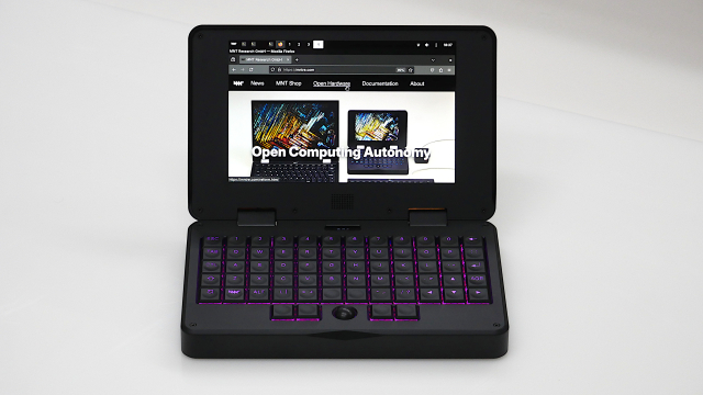 front view of a small (roughly A5 size) black clamshell laptop with ortholinear mechanical keyboard, pink backlit keys, black mini trackball, firefox with mnt research website on screen
