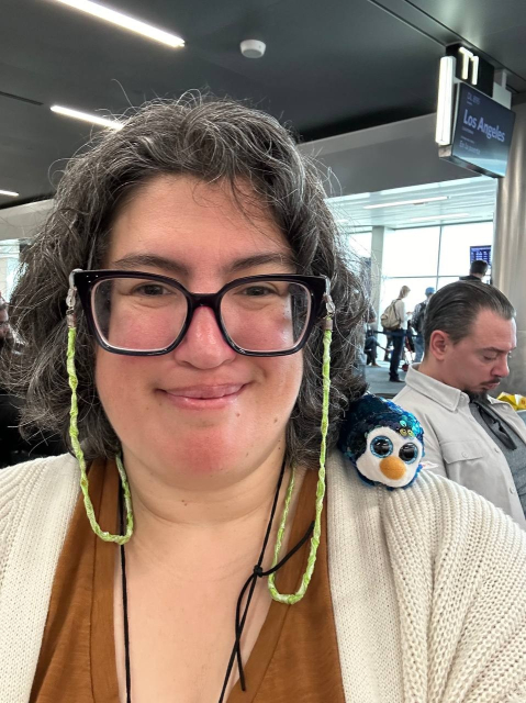 A middle aged person with wavy salt and pepper in a short haircut and oversize glasses smiles at the camera. They are wearing a dark brown shirt with an off white cardigan, and have a small sequined penguin on their shoulder. In the background there is a sign for a flight to Los Angeles.