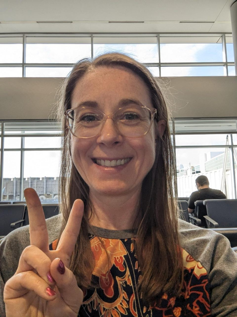 A feminine person with glasses, medium length brown hair, straight except for some curl at the end, and a wide smile sits in an airport, holding up a peace sign with dark red painted nails.