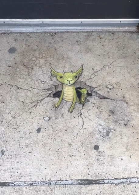 Streetart. Two tiny green dragons with a 3D effect have been painted with chalk on a gray sidewalk in front of a house entrance. The two look out of a painted hole with many cracks in the ground. A large dragon with a triangular head, small black eyes and two beige horns is almost outside, and a smaller dragon is following it. Title:
"This is your annual reminder that not all the little green things that pop up in the spring have flowers."