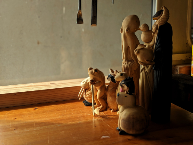 Group of wood carvings : a walrus, a standing up frog, a weird otter, a racoon, an humanoid octopus wearing a dress, a dragon standing with a duck on its head, a gecko, a humanoid monster with a rabbit on its hand and a red robin on its head, a capybara with horns