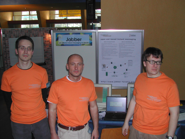 Three dudes standing in front of a poster and a table with some laptops and monitors. All wearing orange t-shirts.
