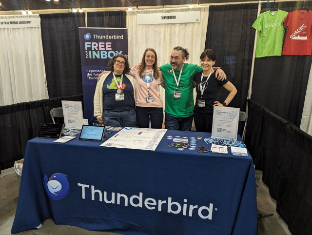 Four people are standing at a Thunderbird booth behind a table, with a stand-up banner that says "Thunderbird - Free the Inbox' visible behind them. The table has two tablets with K-9 mail, stickers, and a informational poster.