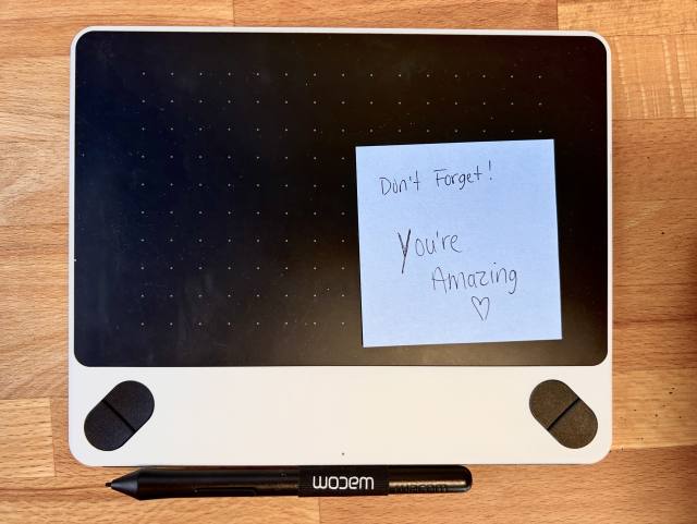 A Wacom drawing tablet with a light blue sticky note that says “Don’t Forget! You’re Amazing ❤️”