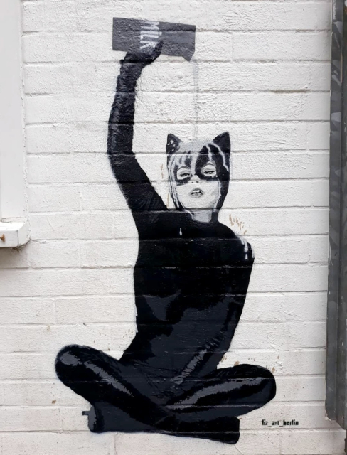 Streetartwall. A mural of a woman in a black full-body suit with cat ears was sprayed onto a white house wall. The cat woman, painted in various shades of black and gray, is sitting cross-legged. She is pouring a carton of milk over herself, which is then running down her head. The mural was applied in several layers using stencils.
Info: The Berlin artist has been on the road with her stencil works and paste-ups since 2016. She applies her motifs to paper and canvas using hand-cut stencils. Liz_art_berlin has an international network and her works can be found in numerous European cities. Her trademark is almost always a small drawn crown.