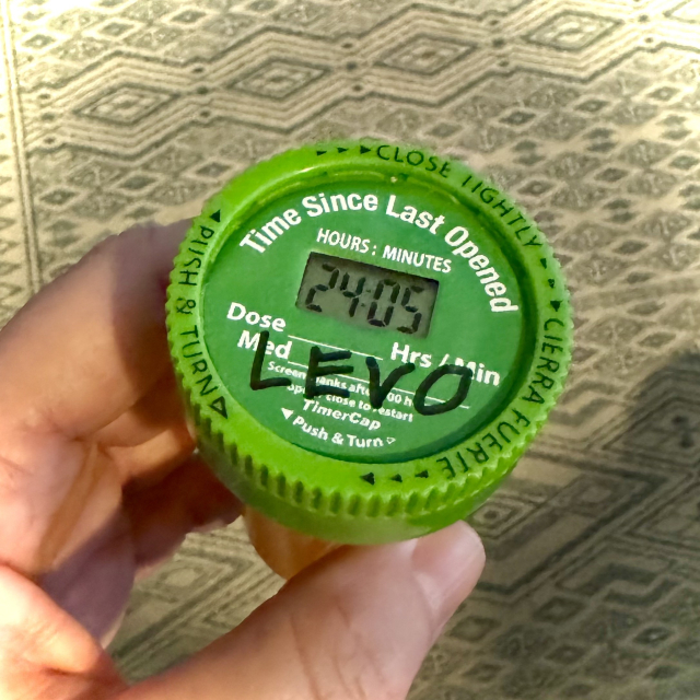 Closeup of my hand holding a pharmacy pill bottle showing the green cap with a cheapo digital count-up timer embedded in it.