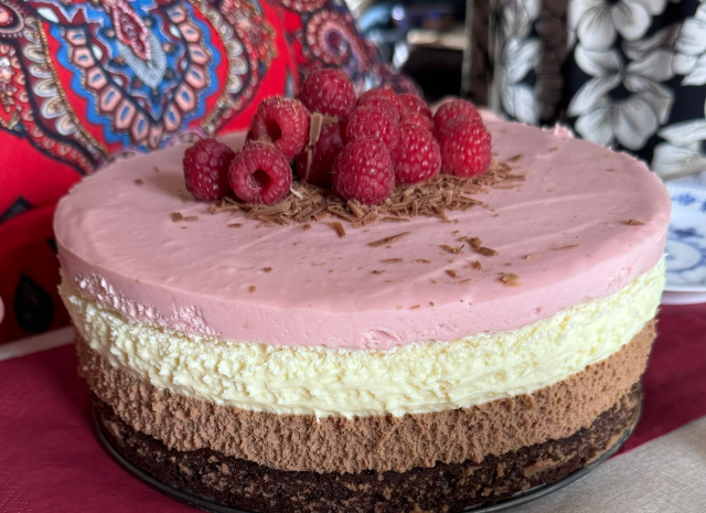 The image shows a layered dessert, possibly a cake, with distinct layers of different colors and textures. The bottom layer is a dark brown, resembling a dense chocolate cake or brownie. Above it is a lighter brown layer, which could be a mousse or cream with a fluffy texture. The next layer is a pale yellow or off-white, suggesting a vanilla or cheesecake layer. The top layer is a soft pink, smooth in texture, possibly a raspberry or strawberry flavored cream. The cake is topped with a handful of fresh raspberries and some chocolate shavings. It's placed on a plate with a blue and white pattern, and in the background, there's a colorful cloth with a red and blue paisley design and a black and white floral pattern.