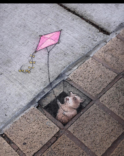 Streetart. A small raccoon with a flying kite was drawn with chalk on a gray sidewalk. A hole with a 3D effect was drawn on one of the darker kerbs, from which the raccoon is now peering out. He is holding the string of a pink kite above him with both hands.
Title: "Larry prefers his outdoor activities as indoors as he can make them."