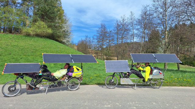 Two solar recumbent bikes with riders parked in front of green meadow, trees and blue sky.