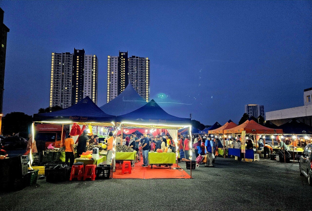 Against the backdrop of a dark blue dawn sky, a brightly lit tent stands out in a field of tents. We are at the pre-Eid farmer's market, and the tent in focus has a long queue if people patiently waiting to pay for their goods (of which you can vaguely see the stacks of lemang in their bamboo casings to the left of the tent, while the queue forms a chain to the right). The scene is full of bright colours of orange, yellow and blue, from the colourful tarp that makes up the tents and table covers