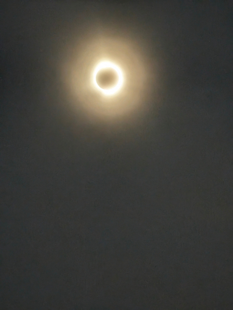 Blurry photo of a total eclipse through cloud.The moon is a dark circle in the middle, surrounded by a bright ring of light, them a second, slightly-dimmer ring.