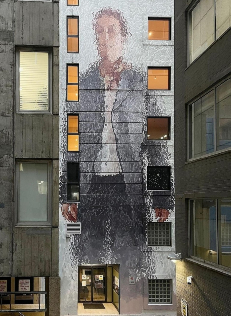 Streetartwall. A mural depicting a historical figure from Sydney was sprayed onto the exterior wall of a narrow high-rise building over approx. 5 floors. Between tall modern buildings, a standing man in an old-fashioned grey suit and white shirt can be seen out of focus. The mural appears gray-metallic and blurred, as if behind frosted glass, and the figure can only be guessed at. This type of painting is the trademark of the artist Fintan Margee.
Info: The mural depicts the first elected Mayor of Sydney, John Hosking (1806-1882) who the lane is named after.