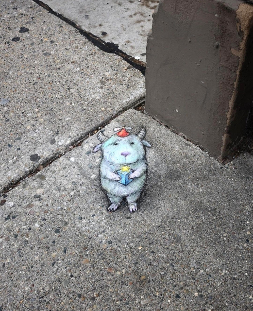 Streetart. A small monster has been painted with chalk and a 3D effect on a sidewalk made of gray stone slabs. The little monster looks like an upright goat with small horns and fluffy white fur. It is wearing a red party hat and is holding a blue gift package with a yellow bow in its hands and looking at us.
Title: "Clarence places small pebbles in festive boxes so they can feel special for a while."