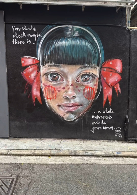 Streetartwall. A beautiful mural of a girl with a message was sprayed on a black street wall. The finely drawn face of a girl with big eyes, short black hair and two red bows on the right and left side looks out at the viewer. Small red stars adorn her nose and her cheeks are painted with two implied planets. Next to her is the text: "You should look: Maybe there's a whole universe in your head."
Info: Hera has sprayed three murals in Hong Kong and this small one was a nice finishing touch before her departure.
