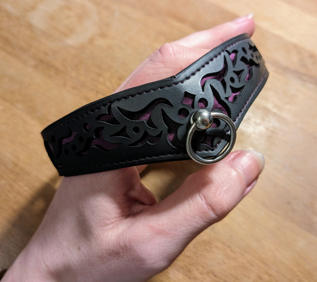 Foto meines neuen Penumbra-Halsbandes, in schwarz (veganes Leder), inkl. Muster und Naht in dunkellila, sowie einem silbernen Ring. Die Aufnahme ist dabei leicht seitlich, nicht frontal.

Im Hintergrund ist noch meine Hand zu sehen, die das Halsband hält, sowie mein Holztisch.